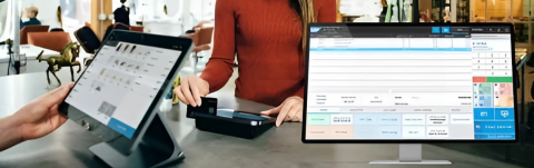 Retail store staff assisting a customer at a counter using LS Retail POS on a tablet and a desktop monitor for transactions.