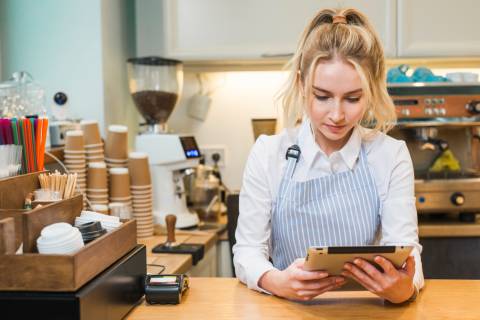  young blonde barista in a light blue striped apron and white shirt stands behind a café counter, focused on a tablet possibly testing one of the best cafe POS systems in the UK. Coffee cups and brewing equipment fill the background.