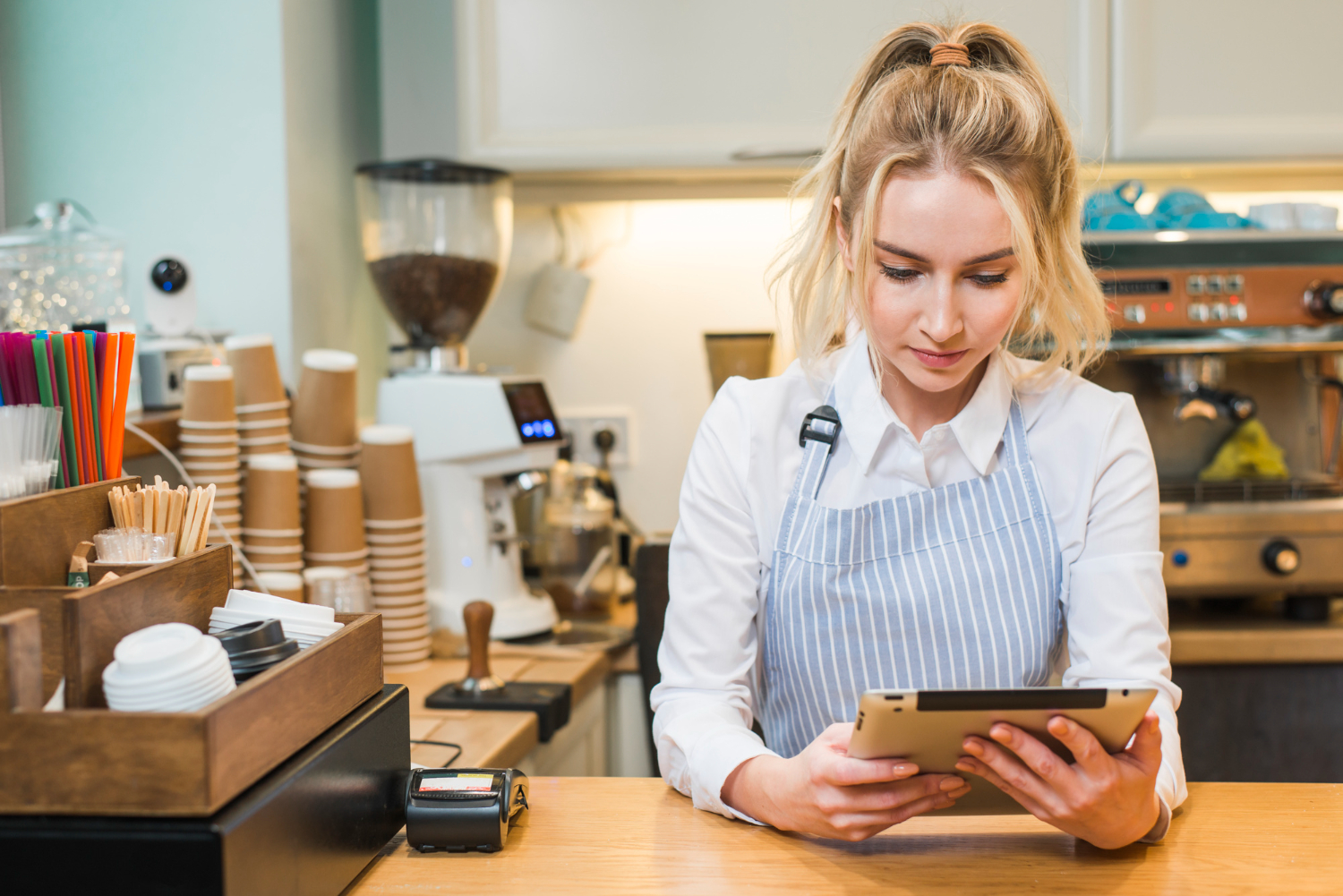 young blonde barista in a light blue striped apron and white shirt stands behind a café counter, focused on a tablet possibly testing one of the best cafe POS systems in the UK. Coffee cups and brewing equipment fill the background.