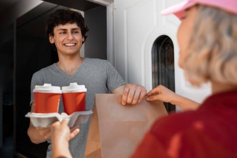  A smiling young man in a grey t-shirt receives a delivery from a person in a pink cap, holding a brown paper bag and a tray with two coffee cups.